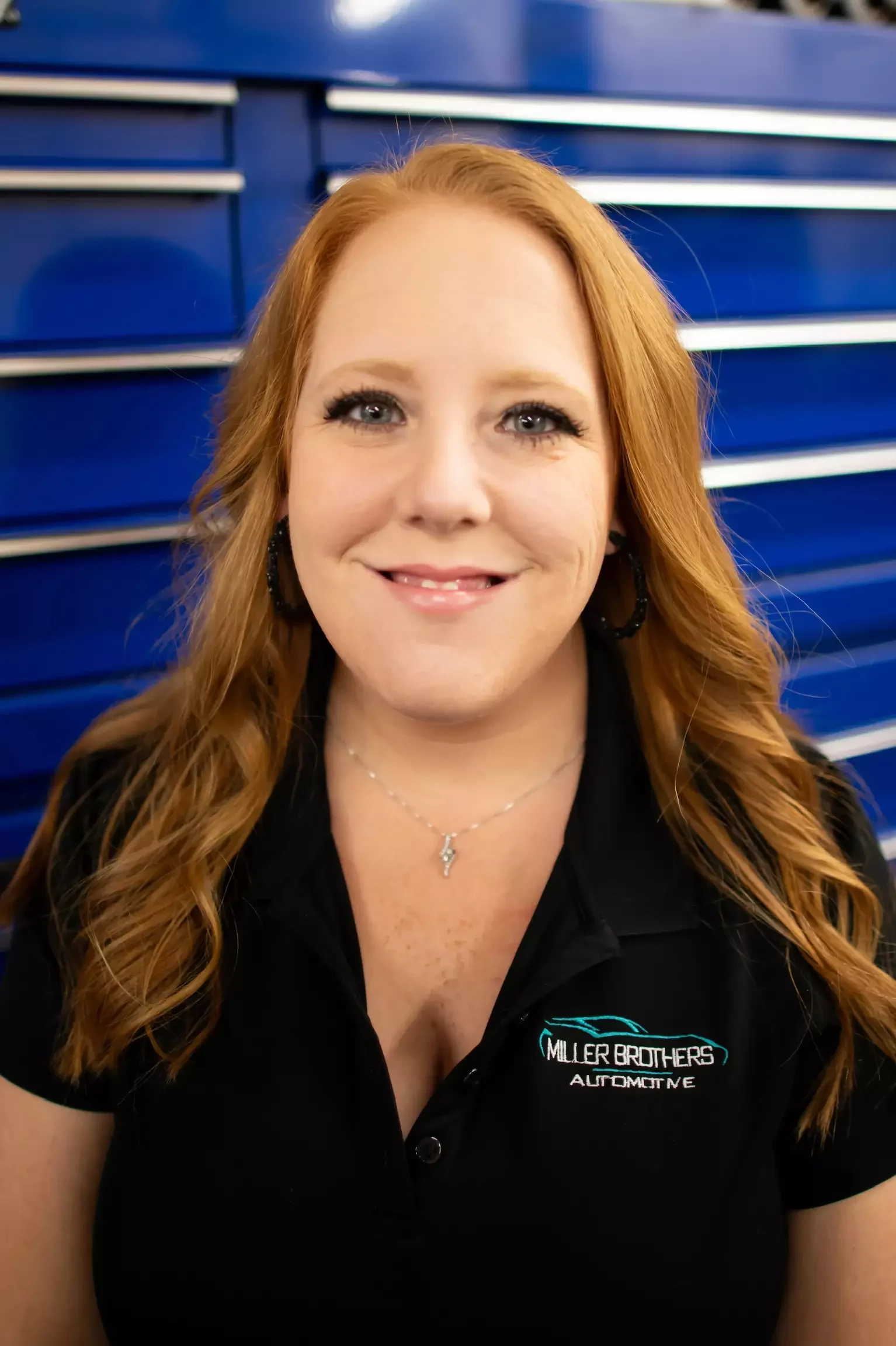 Brandy, Office Manager and Marketing lead at Miller Brothers Automotive, in branded black polo, standing in front of blue tool cabinets. Auto Repair Office Manager and Marketing lead at Miller Brothers Automotive
