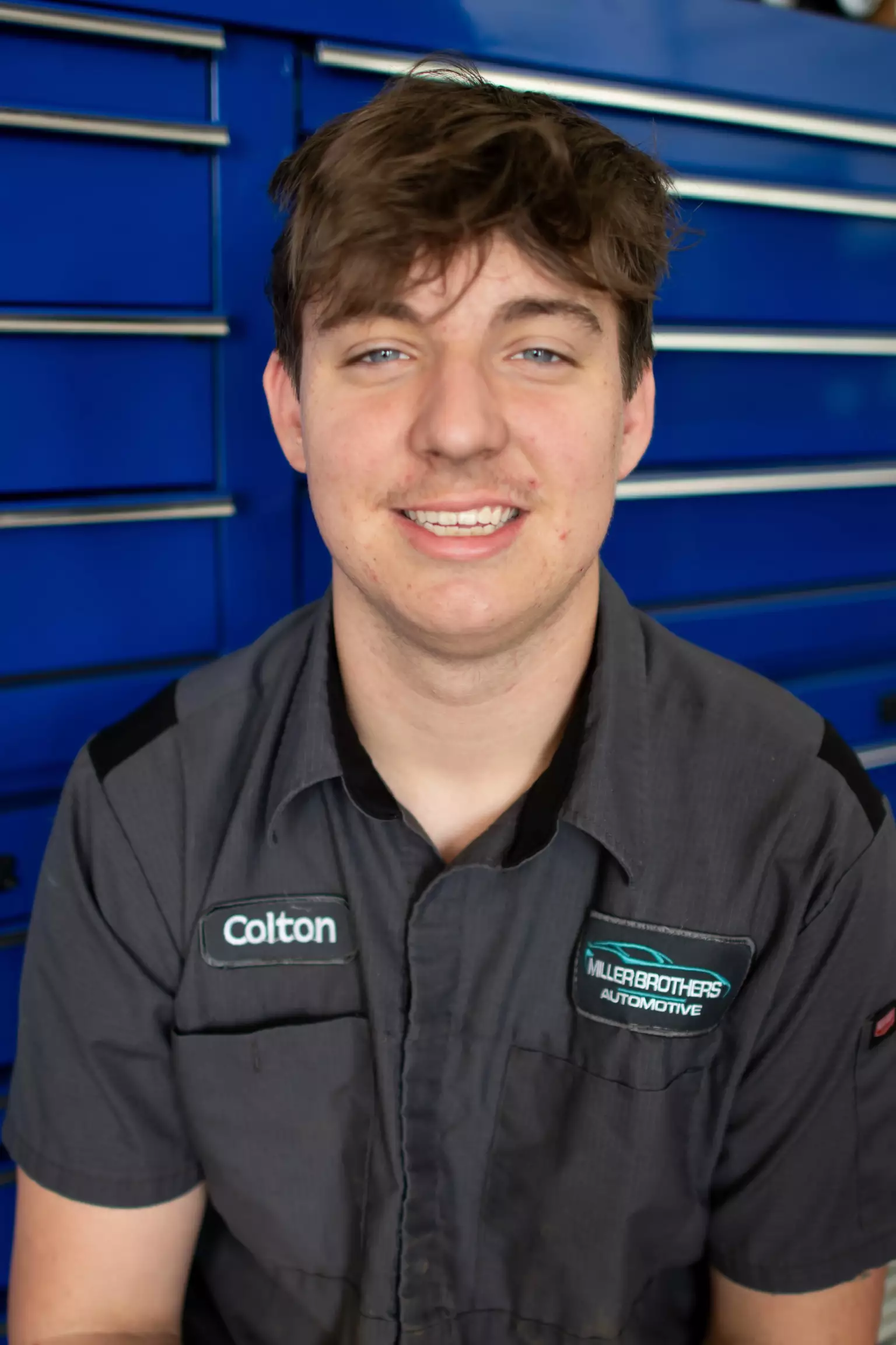 Colton, Technician Apprentice at Miller Brothers Automotive, in gray uniform with name patch, standing in front of blue tool drawers. Auto Repair Technician in Tecumseh OK At Miller Brothers Automotive