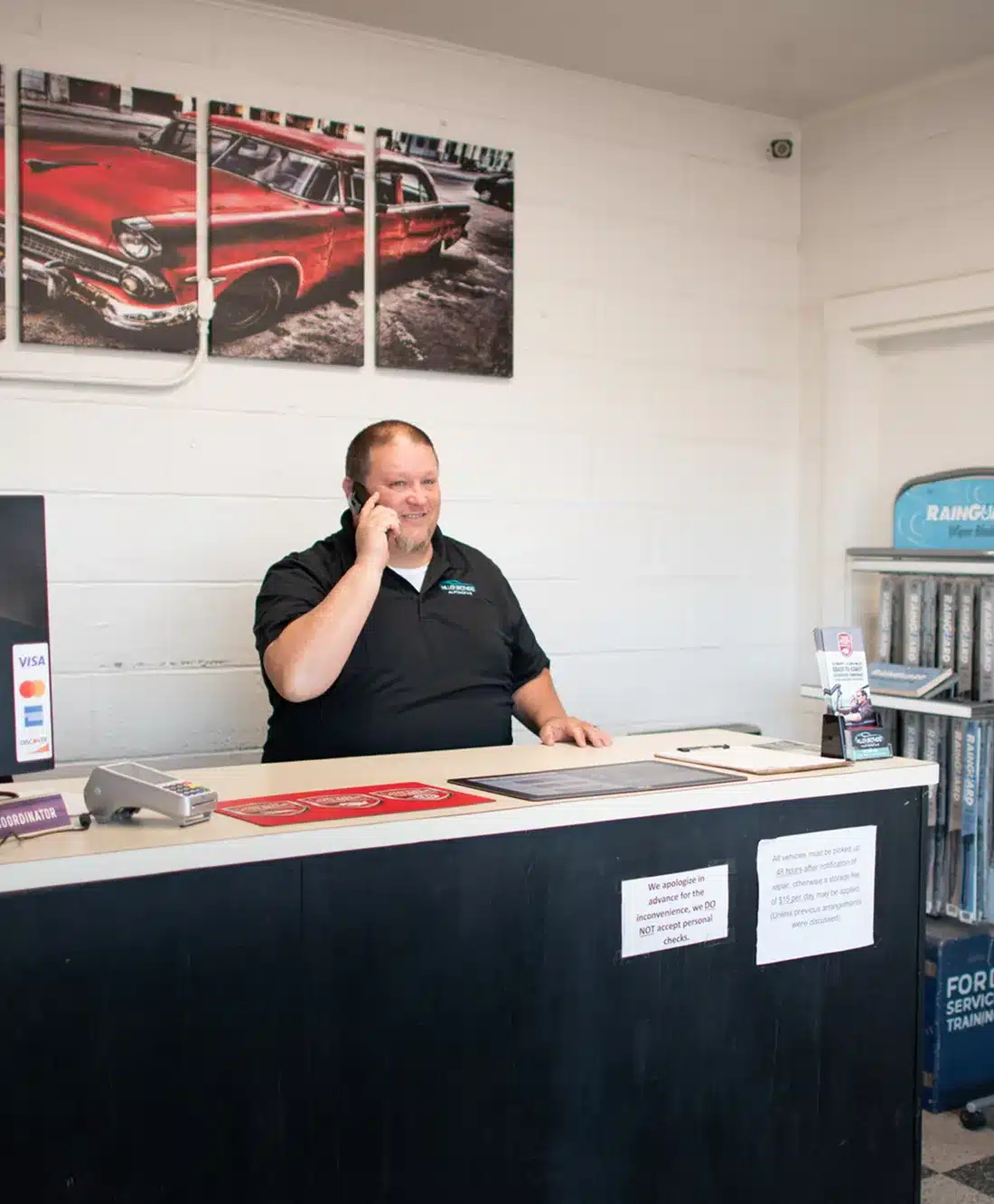 A man in a black shirt stands behind a counter, talking on the phone in an office or service shop. Behind him is a wall with a large photo of a red vintage car. Signs and papers are visible on the counter and wall.