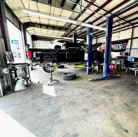 A black pickup truck is lifted on a hydraulic car lift inside Miller Brothers Automotive auto repair shop in Tecumseh, OK, with tools, equipment, and a tire on the floor. A mechanic works beneath the truck. The shop is open, with daylight coming in.