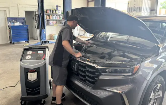 A mechanic inspects the engine of a gray Honda SUV with its hood open inside an auto repair shop, next to a diagnostic machine.