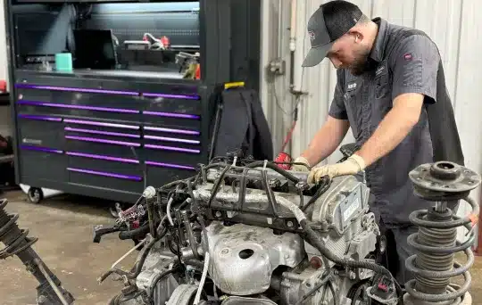 A mechanic wearing gloves and a cap works on a disassembled car engine in a workshop, with a large tool chest and various equipment in the background.