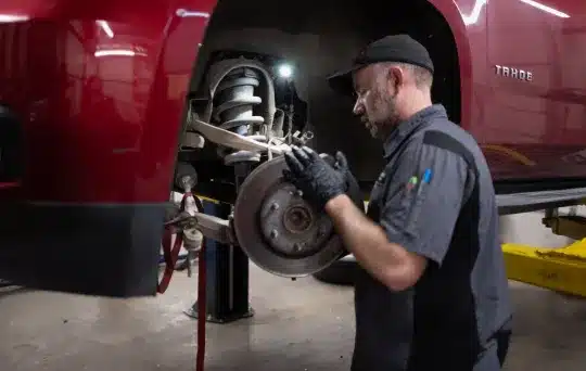 A mechanic wearing gloves and a cap works on the exposed brakes and suspension of a red Chevrolet Tahoe, which is lifted on a car lift in a garage.
