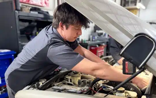 A mechanic in a gray uniform leans over an open car hood, working on the engine inside a well-lit auto repair shop. Tools and equipment are visible in the background.