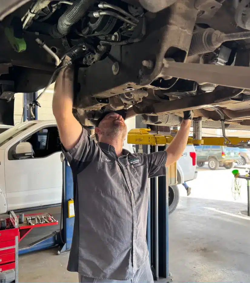 A mechanic in a gray uniform works under a raised vehicle in an auto repair shop, inspecting and repairing components. Tools and other vehicles are visible in the background.