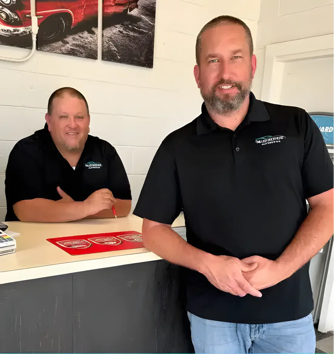Two men wearing black polo shirts stand inside an office. One man stands in front of a counter, and the other sits behind it. Both are smiling. A red car picture is on the wall behind them.