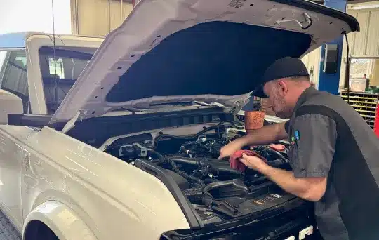 An auto mechanic wearing a cap works under the hood of a white SUV inside a garage, using tools to inspect or repair the engine.