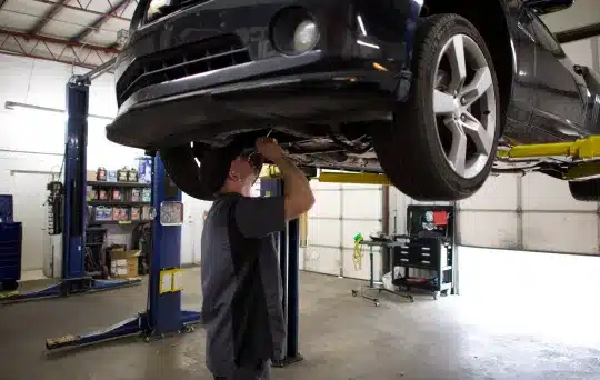 A mechanic works underneath a car that is raised on a hydraulic lift in a garage, with tools and supplies visible in the background.