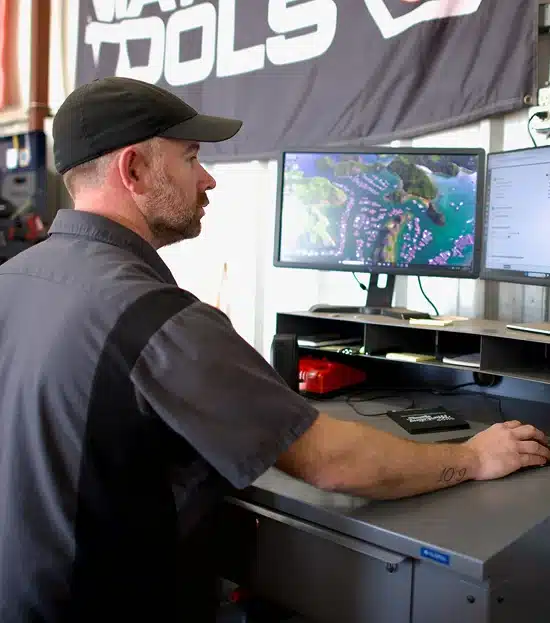 A man in a work shirt and cap uses a desktop computer, handling tasks related to preventative maintenance and auto repair in Tecumseh, OK at Miller Brothers Automotive.