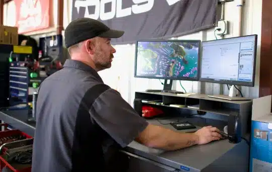 A man in a black cap and work shirt uses a computer with dual monitors at a workstation in a garage or workshop, with tools and equipment visible in the background.