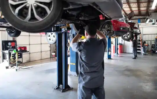 A mechanic works underneath a car lifted on a hydraulic lift in an auto repair shop, with another car and mechanic visible in the background. The workshop is spacious and well-lit.