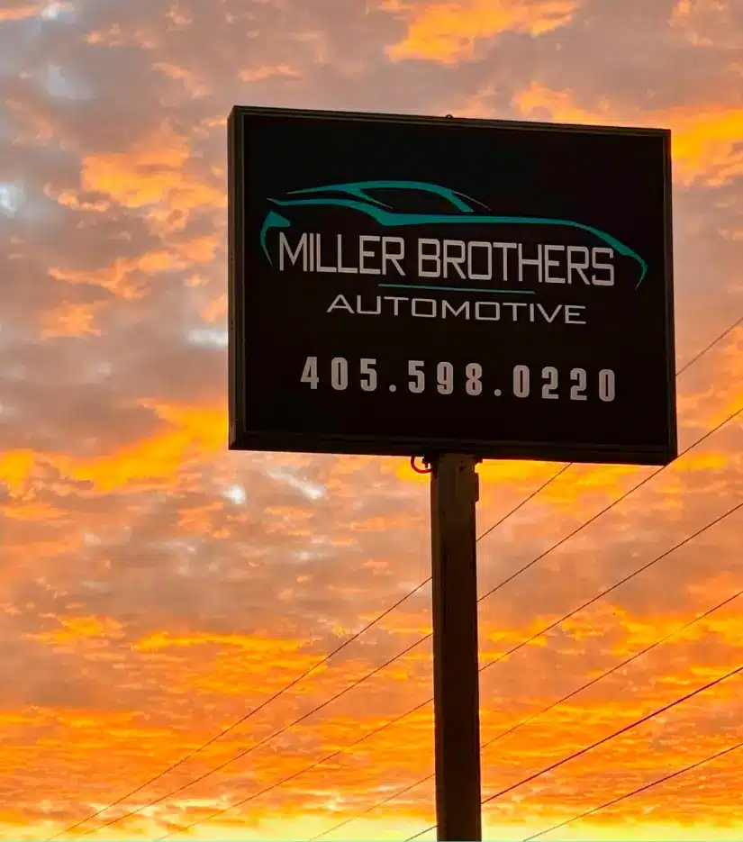 about us, auto repair in Tecumseh, OK at Miller Brothers Automotive. A sign reading Miller Brothers Automotive 405.598.0220 is silhouetted against a vibrant orange and yellow sunset sky with scattered clouds.