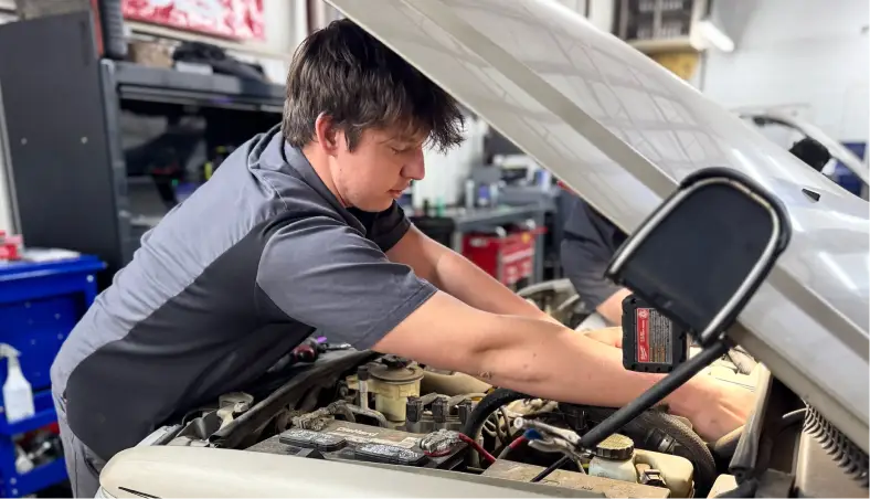 A mechanic wearing a gray uniform performs auto electrical repair under the hood of a car at Miller Brothers Automotive, a trusted provider of comprehensive auto repair in Tecumseh, OK.