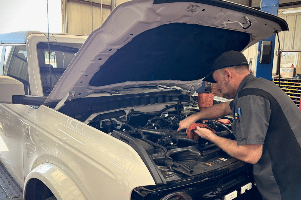 Reliable auto repair. Auto repair shop in Tecumseh, OK. Miller Brothers Automotive. A mechanic wearing a black cap and uniform works under the open hood of a white SUV inside an auto repair shop. Tools and equipment are visible in the background. Engine Repair In Tecumseh, OK At Miller Brothers Automotive