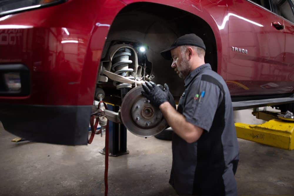 Brake auto repair shop in Tecumseh, OK. Miller Brothers Automotive. A mechanic wearing gloves works on the front brake assembly of a red Chevrolet Tahoe lifted on a car hoist inside an auto repair shop.